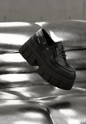 Schwarzer klobiger Plateauschuh mit grobem Profil und Schnürsenkeln, schwebend vor einem glänzenden, metallisch gepolsterten Sofa-Hintergrund dargestellt.