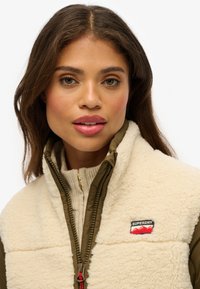 Gilet in pile crema sopra una maglia zip olive, con un'etichetta del logo Superdry e una superficie testurizzata, caratterizzato da un dettaglio della cerniera rossa.