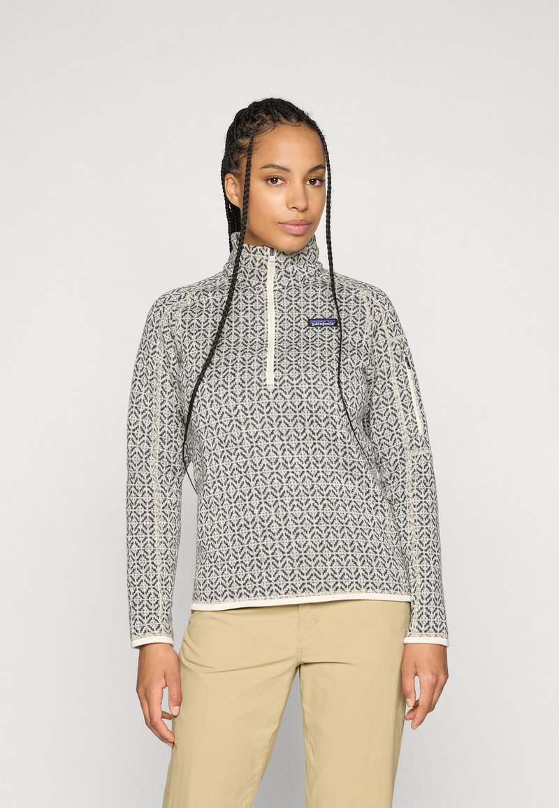Fleece pullover met een geometrisch zwart-wit patroon, hoge kraag, lange mouwen en een ritssluiting aan de voorkant. Heeft crèmekleurige accenten.