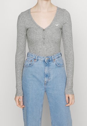 Personne portant un haut gris côtelé à manches longues ajusté avec des boutons et un jean bleu clair taille haute, debout devant un fond clair uni.