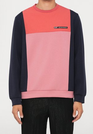 Homme portant un sweat-shirt à manches longues color-block corail, rose et marine, avec un petit logo EA7 Emporio Armani sur la poitrine.