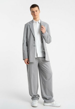 Młody mężczyzna stojący, ubrany w luźny jasnoszary garnitur, białą koszulę i białe trampki na jednolitym białym tle.