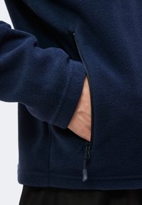 Χέρι μερικώς μέσα στην τσέπη με φερμουάρ στο πλάι ενός σκούρου μπλε fleece μπουφάν, που δείχνει την υφή του υφάσματος και τη λεπτομέρεια του φερμουάρ.