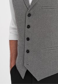 Grå mönstrad väst med en strukturerad tygkvalitet, med en skarp krage, sex svarta knappar och en enkel ficka på vänster sida.