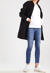 Abrigo ligero negro sobre un sudadera blanca, jeans ajustados azules y zapatillas blancas. El abrigo tiene cierre de botón y bolsillos.