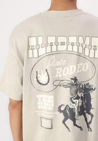 Camiseta de color beige claro con una gran impresión gráfica que representa a un vaquero, un caballo y el texto "ALABAMA State RODEO" en negro.