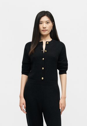 Νέα γυναίκα με μακριά σκούρα μαλλιά, που φοράει μαύρη ριμπ πλεκτή ζακέτα με χρυσά κουμπιά και μαύρο ριμπ πλεκτό παντελόνι, πάνω σε μονόχρωμο φόντο.