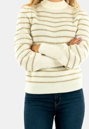 Osoba z założonymi ramionami, ubrana w kremowy sweter z brązowymi poziomymi paskami i ciemnoniebieskie dżinsy, na jednolitym tle.