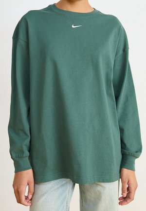 Henkilö, jolla on yllään löysä, tummanvihreä pitkähiha-Nike-paita ja vaaleansiniset farkut, kädet rentoina sivuilla, näkyvissä yläruumis ja reidet.