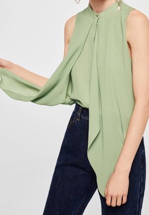 Zielona bluzka bez rękawów z wysokim kołnierzem i długim asymetrycznym przodem w tandemie z ciemnoniebieskimi jeansami na osobie.