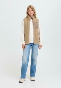 Jeune femme portant un gilet en polaire beige par-dessus un pull blanc, un jean bleu et des baskets claires, se tenant devant un fond clair et uni.
