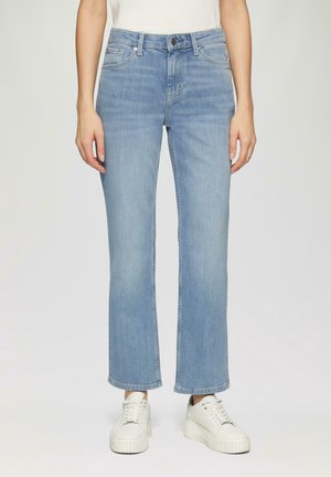 Femme portant un jean droit bleu clair et des baskets blanches à plateforme, debout devant un fond uni.