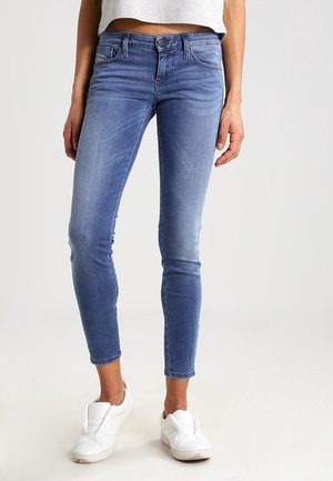 Jean bleu slim porté par une personne avec des baskets blanches slip-on et un top court gris clair sur un fond uni.