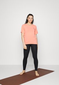 T-shirt cor-de-coral de manga curta com um logótipo, combinada com leggings pretas. A modelo está descalça sobre um tapete castanho, em frente a um fundo neutro.