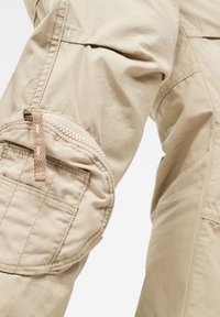 Detailní záběr béžových cargo kalhot, který ukazuje zipovou kapsu a prošívání na klopě kapsy.