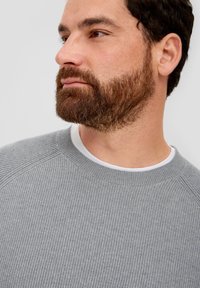 Gray ribbed sweater with a round neckline, worn over a white shirt. Features raglan sleeves and a textured knit design.