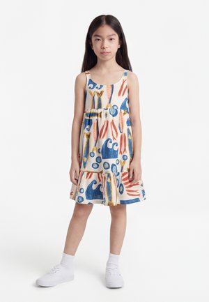 Fiatal lány áll, ujjatlan fehér ruhát visel, amelyen kék, narancs és sárga absztrakt hal- és hullámminták vannak, fehér zoknit és fehér tornacipőt visel.