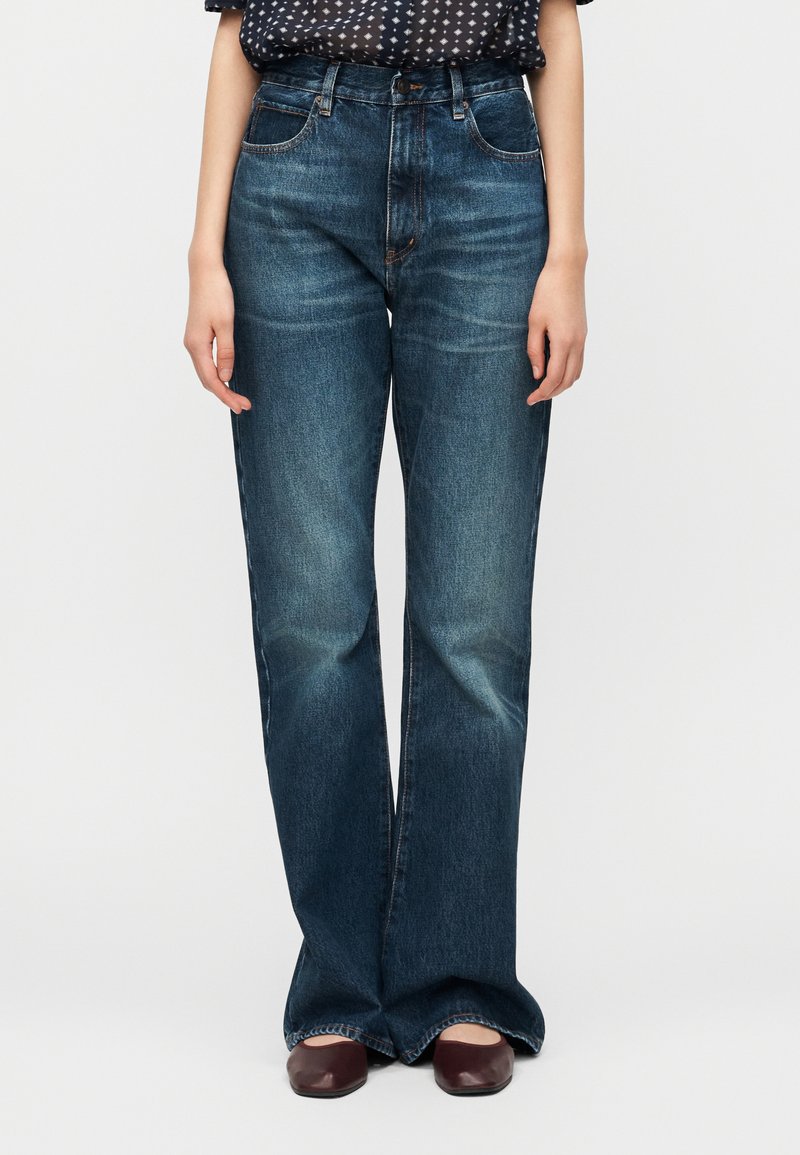 Γυναίκα που φορά υψηλόμεσα, σκουρό μπλε τζιν με ίσια παντελόνια και μπορντό κλειστά παπούτσια, στέκεται με τα χέρια άνετα στο πλάι.