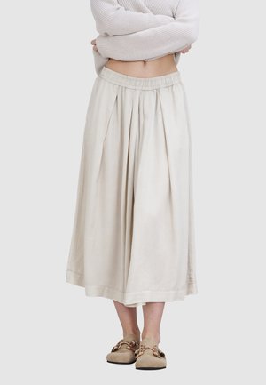 Jupe beige, longueur genou, avec une taille élastique et des plis. Tissu doux au toucher lisse. Associée à des chaussures beige clair avec des accents dorés.