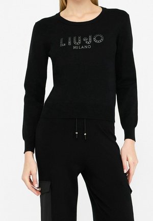 Frau trägt schwarzen Langarm-Pullover mit "Liu Jo Milano" Schriftzug aus Nieten und passende schwarze Hose mit seitlicher Taschen-Detail.