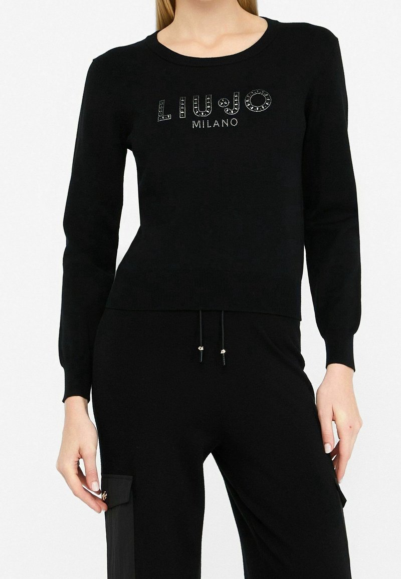 Femme portant un pull noir à manches longues avec le texte « Liu Jo Milano » en clous et un pantalon noir assorti avec un détail de poche latérale.