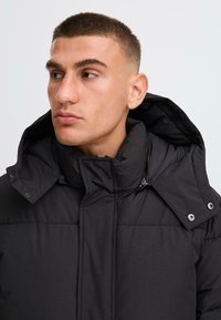 Čierna prešívaná bunda s vysokým golierom, kontrastnými švami a zapínaním na cvočky. Disponuje zipsom a kapucňou.