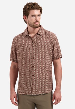 Mann trägt braunes, gemustertes kurzärmeliges Hemd mit Knöpfen und khakifarbene Hose, schaut seitlich vor einfarbigem Hintergrund.