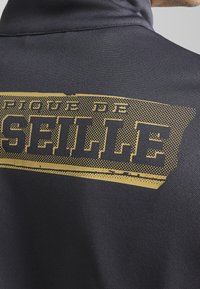 Camisa polo negra con textura de puntos, con un gráfico dorado prominente que dice "PIQUE DE SELLE" en la parte de atrás.