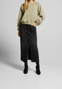 Olivgrön sweatshirt med långa ärmar, matchad med en svart denim-maxikjol med en framficka och fransad nederkant. Svarta åtsittande ankboots.