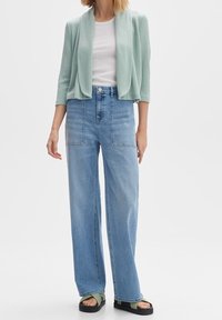 Ljus mintgrön kortärmad cardigan, vit åtsittande topp, högmidjade ljusblå vida jeans och svarta platåsandaler med gröna detaljer.