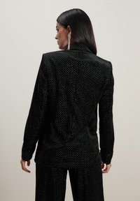Sorte fløjl blazer med hvide prikker, enkeltbrystet design, lange ærmer og struktureret krave. Teksturen ser blød ud.
