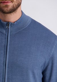 Uomo in maglione blu con zip, colletto a coste e una barba visibile sul volto, in primo piano su uno sfondo semplice.
