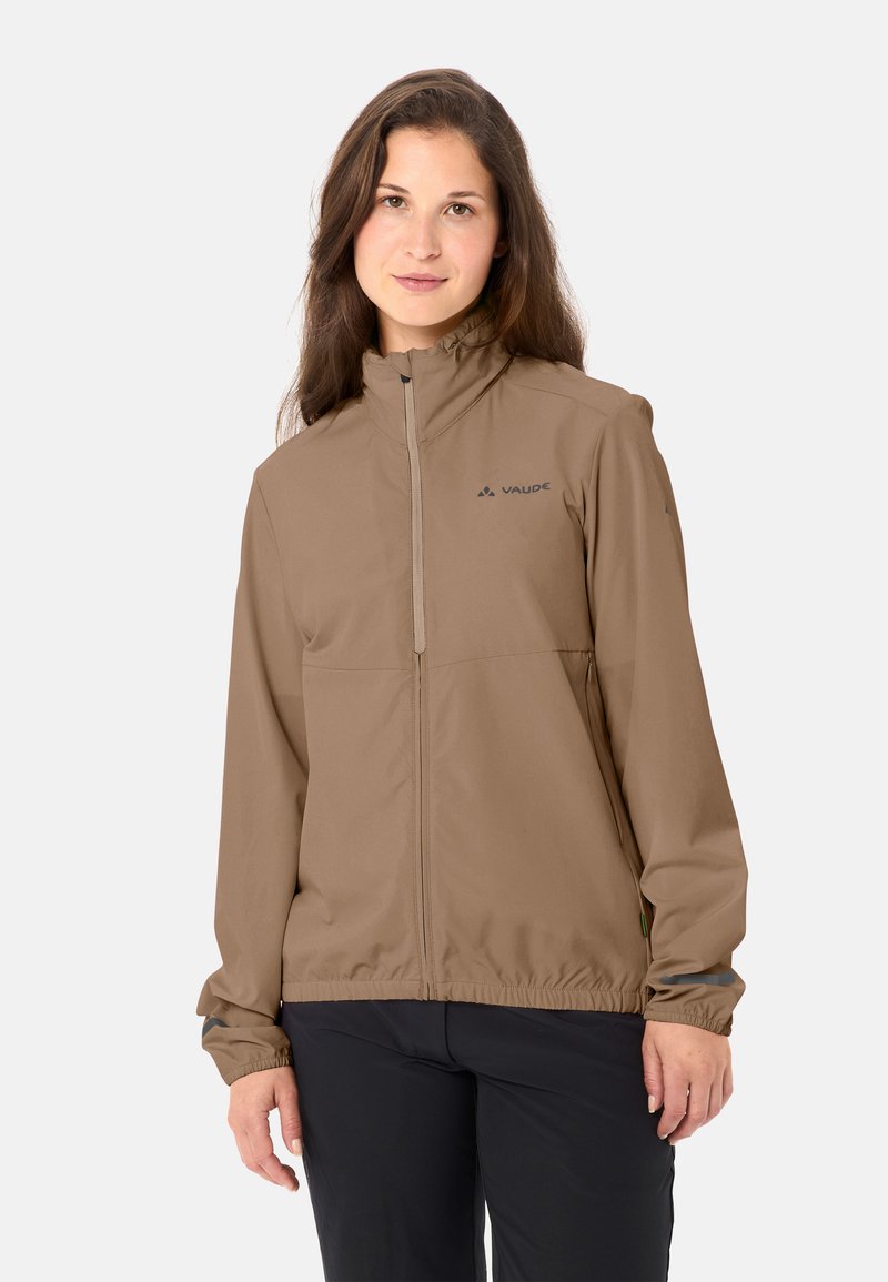 Donna che indossa una giacca marrone chiaro con zip e collo alto e pantaloni neri, in piedi davanti a uno sfondo chiaro e uniforme.