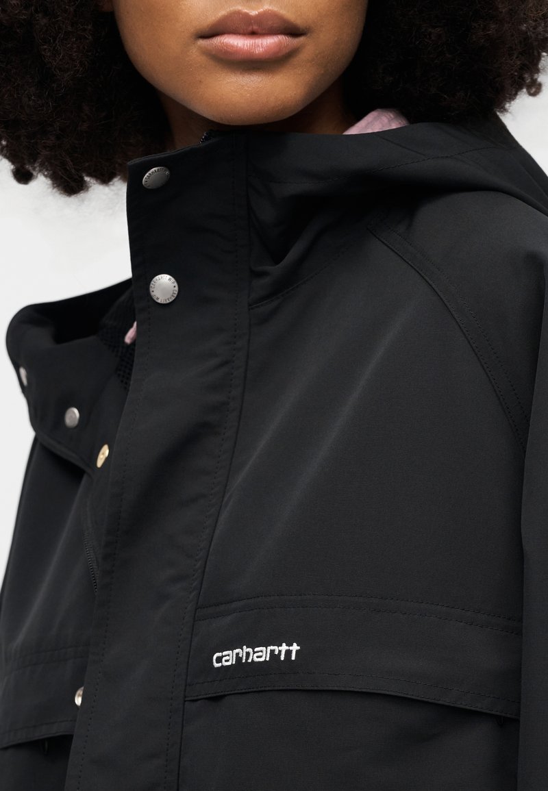 Giacca impermeabile nera con cappuccio, chiusure a scatto e un discreto logo Carhartt bianco sul lato destro inferiore. Tessuto dalla texture liscia.