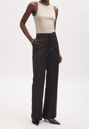 Person wearing sleeveless beige top, high-waisted wide-leg black trousers, and black pointed-toe heels, standing with one hand in pocket.