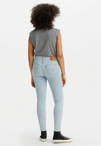 Jeans skinny de color azul claro con tiro alto, que cuentan con bolsillos traseros y una etiqueta de cuero marrón. La modelo lleva una camiseta sin mangas gris y zapatillas negras.