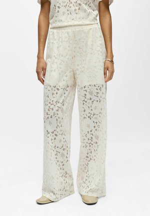 Femme portant un pantalon large en dentelle florale blanche avec une taille élastique, debout devant un fond blanc.