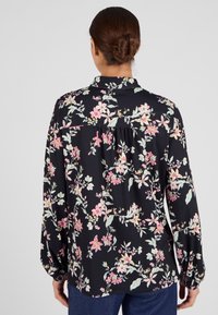 Blusa negra de flores con mangas largas, que presenta patrones de flores en rosa y verde, un collar y un detalle fruncido en la parte de atrás.