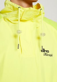 Veste à capuche de couleur jaune fluo avec cordons de serrage et logo noir "ejko Freestyle". Elle présente une texture lisse et des accents verts contrastants.