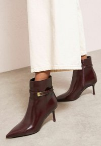 Šauras, padziļinātais potīšu zābaku pāris tumšā burgundijas ādas krāsā ar dekoratīvu sprādzi un rāvējslēdzēju, kā arī plānu vidēja augstuma papēdi.