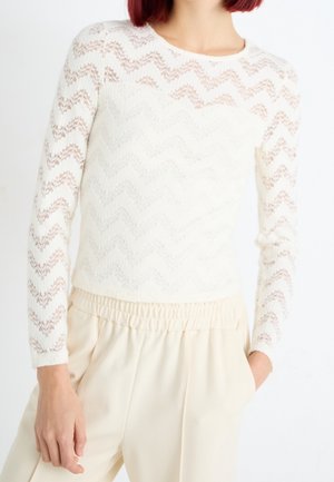 Frau trägt ein weißes langärmliges Oberteil mit Zickzack-Spitze und cremefarbene hoch taillierte Hosen mit elastischem Bund, Hand in der Tasche, vor weißem Hintergrund.