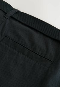 Gros plan sur un pantalon en tissu noir avec une poche passepoilée à l'arrière et une ceinture ou bande texturée noire.