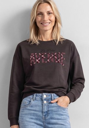 Smilende kvinde med blondt hår iført en mørk sweatshirt med teksten "La Petite Avenue" og lyseblå, højtaljede jeans, hånd i lommen.