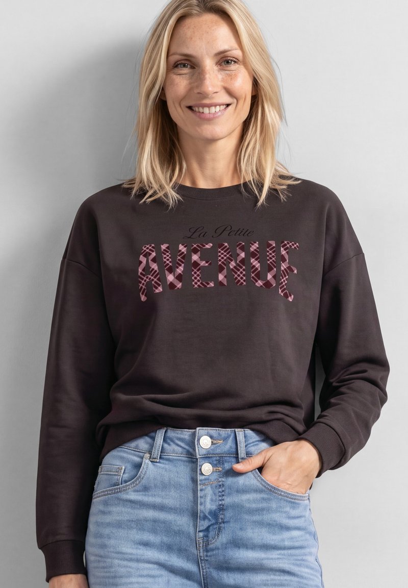 Femme souriante aux cheveux blonds portant un sweat-shirt foncé avec le texte "La Petite Avenue" et un jean taille haute bleu clair, main dans la poche.