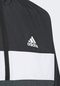 Chaqueta cortavientos en blanco y negro con cierre frontal, que presenta una franja horizontal blanca y un logo bordado de Adidas en el pecho.