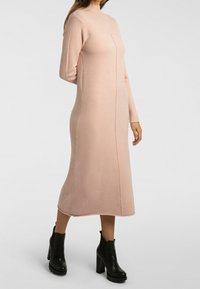 Robe en tricot rose avec un col montant, des manches longues et une coupe ajustée. Présente une couture centrale à l'avant et une longueur mi-mollet. Portée avec des bottines noires à talons carrés.