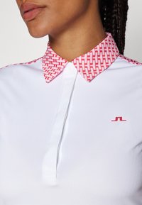 Bílá polokošile s límcem s červeným geometrickým vzorem a malým červeným logem na levé straně hrudi. Hladká textura látky.