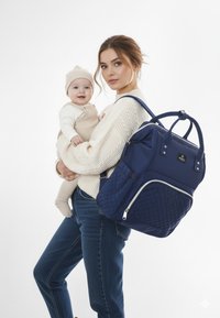 Marinblå, vadderad tygryggsäck med dubbla handtag, dragkedjor och en framficka, bärs av en person som håller ett spädbarn.