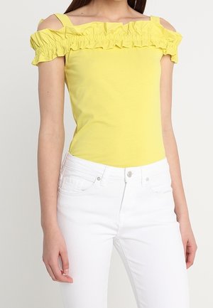Femme portant un haut jaune à volants et épaules dénudées avec des bretelles, associé à un jean blanc taille haute sur un fond uni.