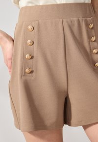 Shorts marron en tissu doux, avec une taille haute et des boutons dorés décoratifs sur le côté, créant un look cintré.